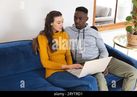 Diverse couple sitting on sofa using laptop at home Stock Photo