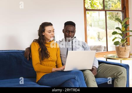 Diverse couple sitting on sofa using laptop at home Stock Photo