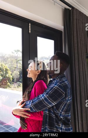 Happy diverse couple embracing and looking through window at home Stock Photo