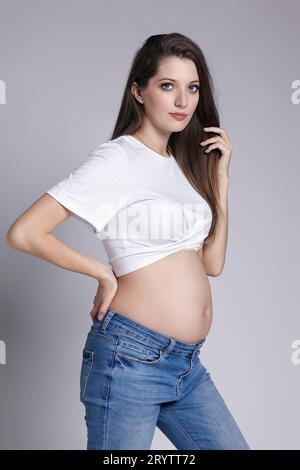 Young pretty pregnant woman in white t-shirt and jeans. on gray background. Belly exposed. Female on 5th month of pregnancy. Stock Photo