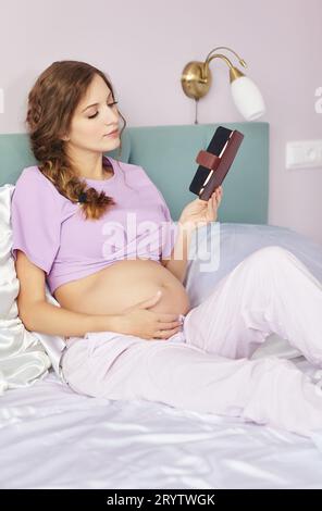 A pregnant woman lies in bed of her domestic bedroom. Female reads an e-book. Stock Photo