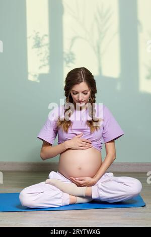 A pregnant woman with long brown hair sits cross-legged, meditating and practicing yoga during pregnancy. Woman looks at her pre Stock Photo