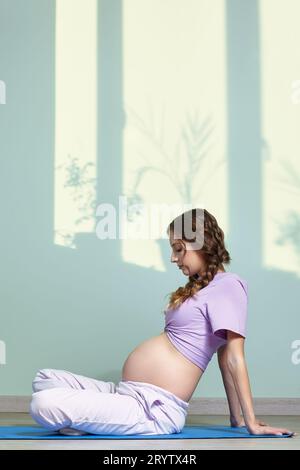 A pregnant woman with long brown hair sits cross-legged, meditating and practicing yoga during pregnancy. Woman looks at her pre Stock Photo