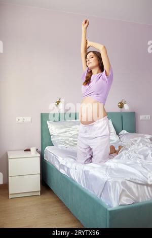 A pregnant woman kneeling on the bed and stretching in bed of her domestic bedroom. Stock Photo