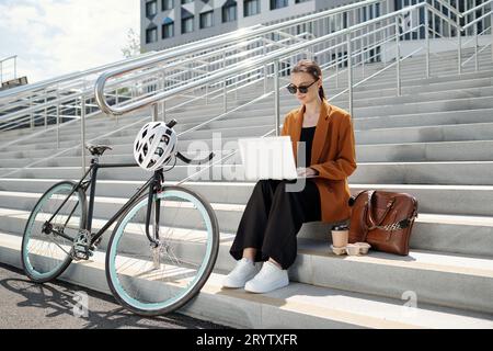 Young serious female freelancer in casualwear using laptop outdoors while sitting on staircase in front of her bicycle with protective helmet Stock Photo