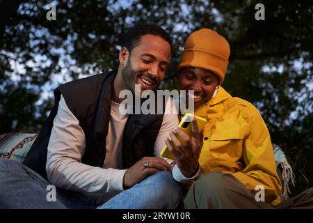 Happy young African American couple using mobile outdoors. Two joyful friends sitting looking phone. Stock Photo