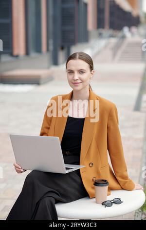 Young successful female student or freelancer in quiet luxury attire looking at camera while sitting in urban environment and using laptop Stock Photo