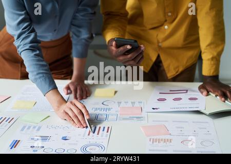 Hand of young businesswoman pointing at financial chart on paper document while explaining it to African American male colleague Stock Photo