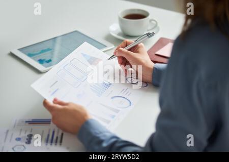 Focus on hand of young female accountant or auditor holding pen over document with financial data while sitting by desk and analyzing charts Stock Photo