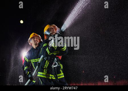 Firefighters using a water hose to eliminate a fire hazard. Team of female and male firemen in dangerous rescue mission. Stock Photo