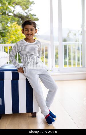 Portrait of happy biracial boy wearing pyjamas in bedroom in front of window with view of garden Stock Photo