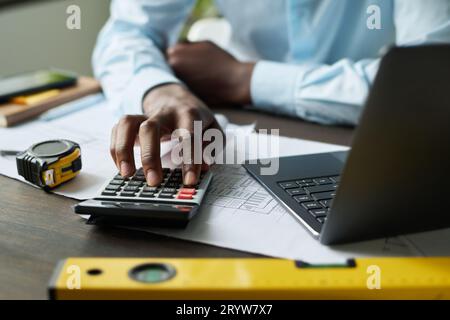 Focus on hand of African American engineer pressing buttons of calculator while sitting by workplace with blueprints and handtools in office Stock Photo