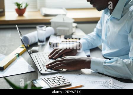 Side view of young male architect sitting by desk with blueprints, handtools, calculator and other stuff and typing on laptop keyboard Stock Photo