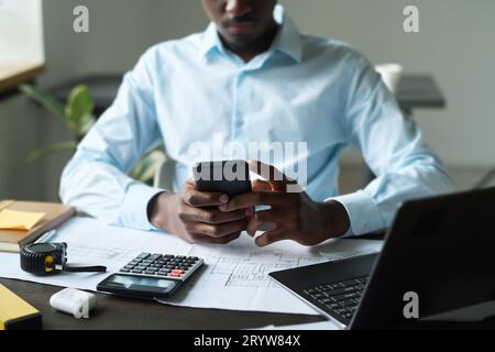 Cropped shot of young African American engineer texting in smartphone while sitting by desk with blueprints in front of laptop in office Stock Photo