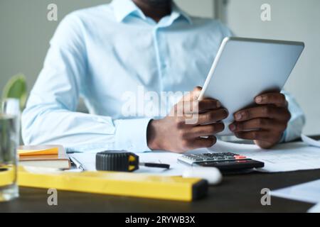 Cropped shot of young African American male engineer holding tablet while sitting by workplace with handtools and sketches and networking Stock Photo