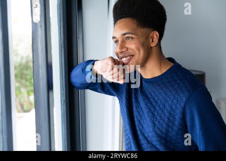Happy biracial man standing in bedroom looking out of window, smiling Stock Photo