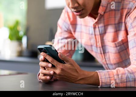Midsection of happy biracial man leaning on countertop using smartphone in sunny kitchen Stock Photo