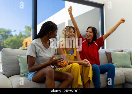 Group of happy diverse female friends sitting on sofa, watching sport and cheering Stock Photo