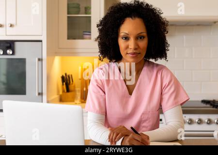 Portrait of biracial female medical worker in kitchen with laptop, working at home Stock Photo