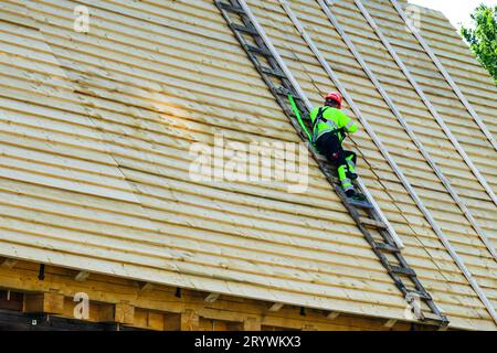 Worker nails new clapboards layer on roof before installing clay tiles, restoration of historic wooden house, blue sky background Stock Photo