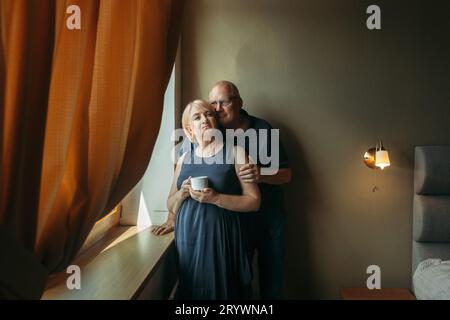 Portrait of a married elderly couple standing at the window. The man hugs the woman by the shoulders. Stock Photo