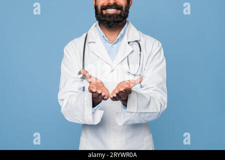 Bearded male medical doctor wearing white medical coat with a stethoscope, holding something invisible in his palms Stock Photo