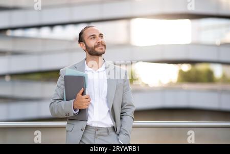 Dreamy Young Businessman With Laptop Standing Outdoors And Looking At Copy Space Stock Photo
