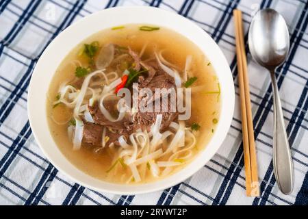 Traditional vietnamese spicy soup Pho Bo with beef piece, rice noodle and herb in plate with chopstick and spoon on checkered tablecloth, closeup. Stock Photo