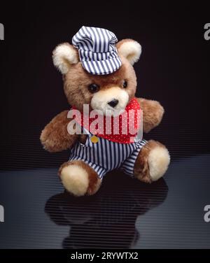 Cute plush teddy bear dressed as a train conductor with striped overalls, cap and red bandana, sitting on reflective surface against dark background Stock Photo