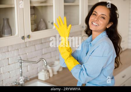 Portrait of joyful woman putting on rubber gloves, ready to tidy house or washing dishes standing in kitchen, free space Stock Photo