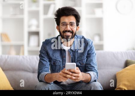 Happy Indian Man With Smartphone In Hands Relaxing On Couch At Home Stock Photo
