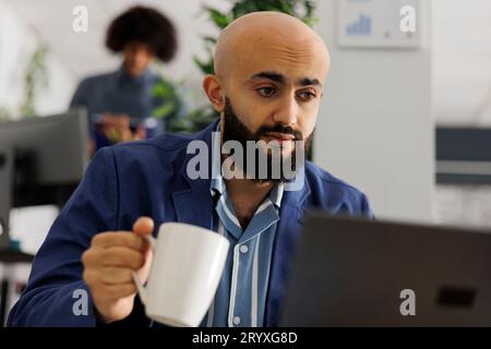 Arab executive manager analyzing market research report on laptop and drinking coffee in business office. Entrepreneur holding tea cup and managing project in coworking space Stock Photo