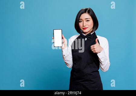 Young asian woman cafe worker presenting smartphone empty screen mockup and showing thumb up gesture. Waitress showcasing mobile phone blank touchscreen and expressing approval sign portrait Stock Photo