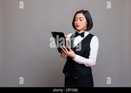 Young asian waitress in uniform checking cafe menu on digital tablet. Catering service staff managing online order and tapping on touchscreen while posing in studio on gray background Stock Photo