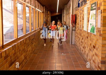 Diverse happy schoolgirls with bags running and embracing in elementary school corridor Stock Photo