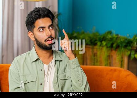 Eureka. Thoughtful clever inspired Indian man make gesture raises finger came up with creative plan feels excited with good idea inspiration motivation, question at home apartment. Guy sits on couch Stock Photo