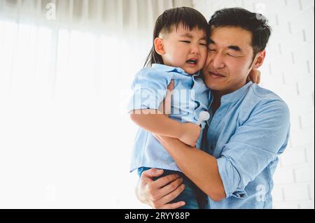Asian Japanese father carrying and consoling his crying son in bedroom at his home with window and white curtain background. Peo Stock Photo