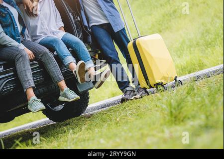 Closeup lower body of group of friends relaxing on SUV car trunk with trolly luggage along road trip with autumn mountain hill b Stock Photo