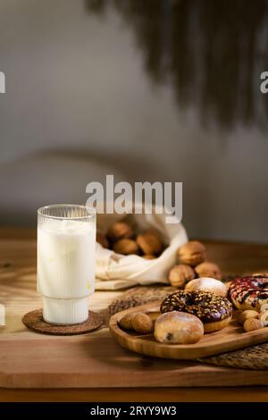 A wooden tray holds three donuts and chocolate-filled cookies with bite-sized biscuits. White fabric cradles walnuts in the background, near a glass o Stock Photo