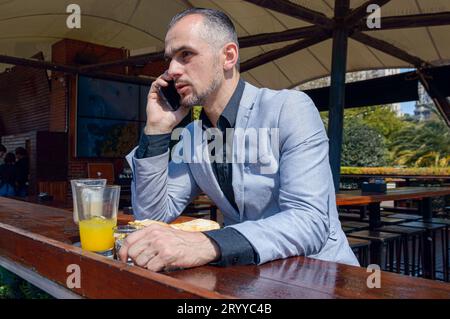 adult Caucasian business man with beard and short hair, concentrated talking on phone call while having breakfast sitting outside restaurant during su Stock Photo