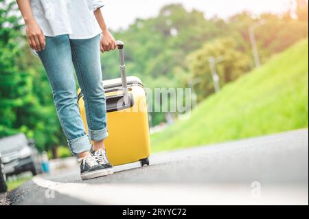 Closeup lower body of woman handling yellow trolly luggage along road trip with mountain hill background. Freedom girl on road w Stock Photo