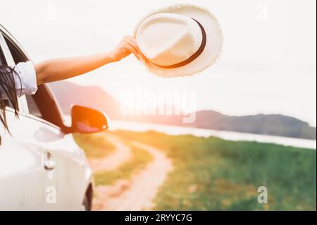 Happy woman hand holding hat outside open window car with meadow and mountain lake background. People lifestyle relaxing as trav Stock Photo