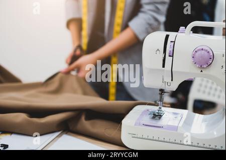 Dressmaker cutting dress fabric on sketch line with sewing machine foreground. Fashion designer tailor or sewer in workshop stud Stock Photo