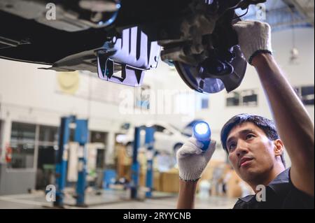 Asian male mechanical hold and shining flashlight to examine car disk brake pad wear of automotive vehicle. Safety suspension in Stock Photo