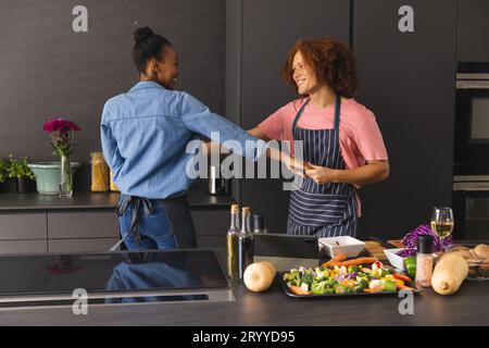 Happy diverse couple in aprons preparing meal having fun dancing in kitchen Stock Photo