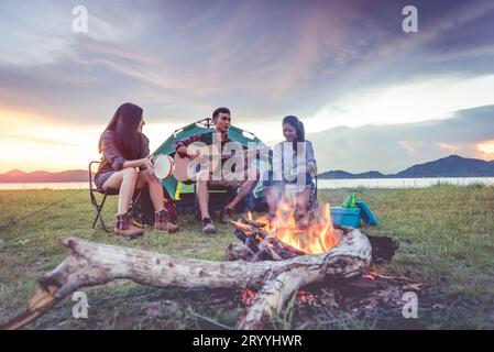 Group of travelers camping and doing picnic and playing music together. Mountain and lake background. People and lifestyle. Outd Stock Photo