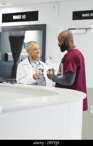 Diverse male and female doctors talking and drinking coffee in reception at hospital Stock Photo