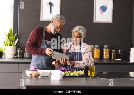 Happy senior biracial couple in aprons using tablet preparing vegetables in kitchen at home Stock Photo