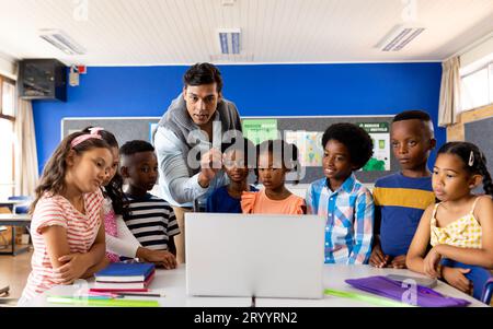 Biracial male teacher using laptop teaching children in elementary school class Stock Photo