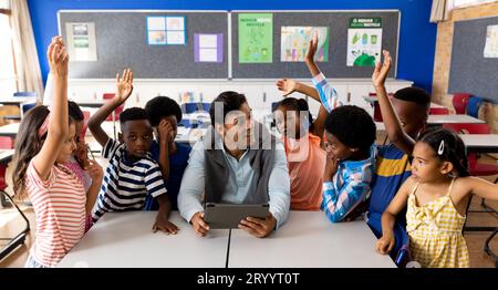 Diverse male teacher using tablet with children raising hands in class at elementary school Stock Photo